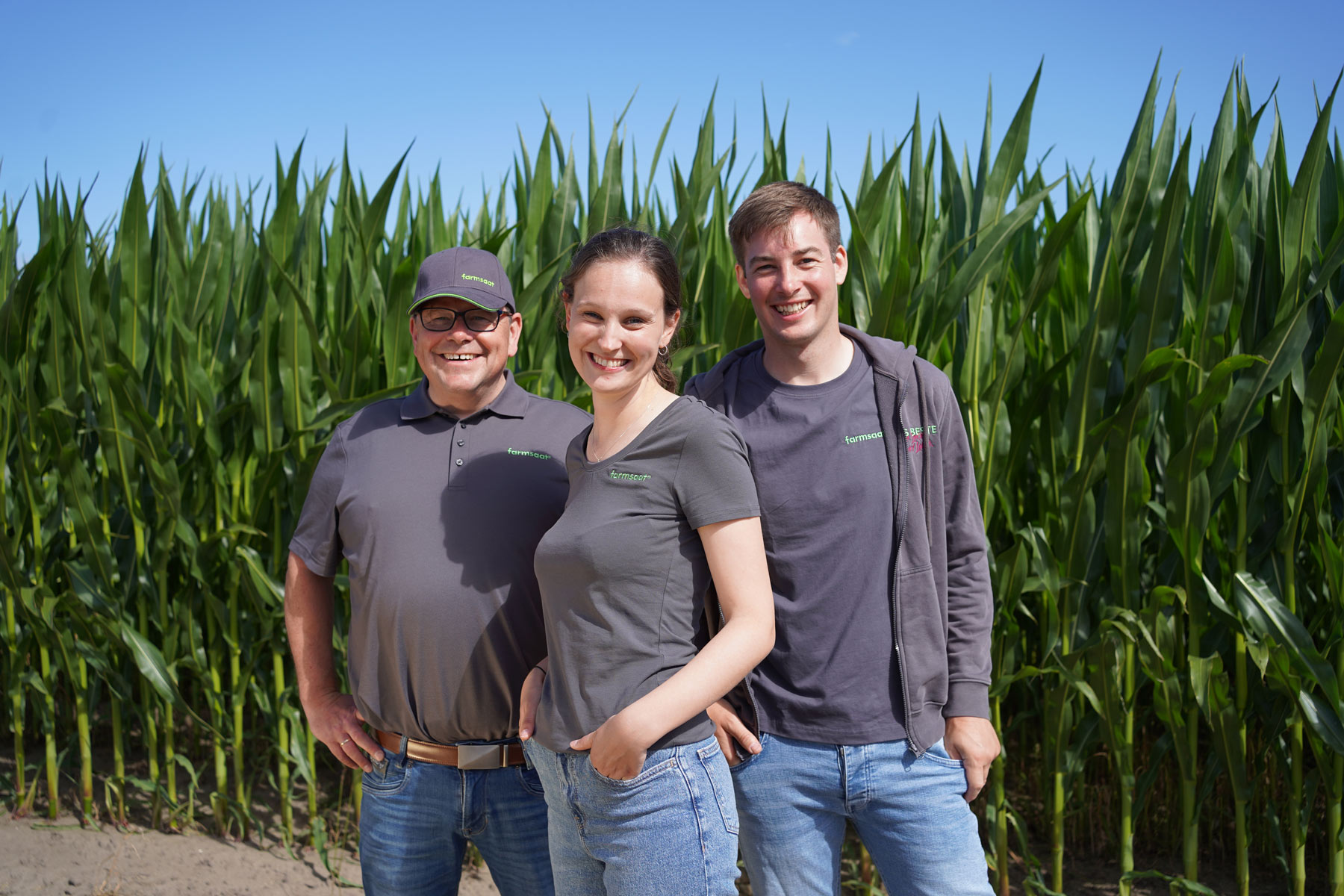 Drei farmsaat-Mitarbeitende, darunter 2 jüngere und ein älterer, stehen lächelnd vor einem Feld